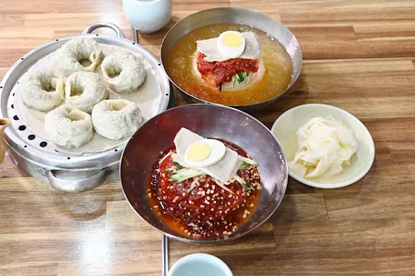 Two bowls of Busan milmyeon noodles topped with egg and red seasoning, beside steamed dumplings